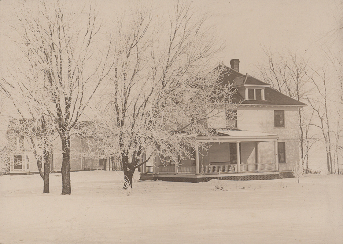 Photograph of Kildee Cottage from the side. It is surrounded by snow on the ground and snowy trees. Norton House is visible in the left background.