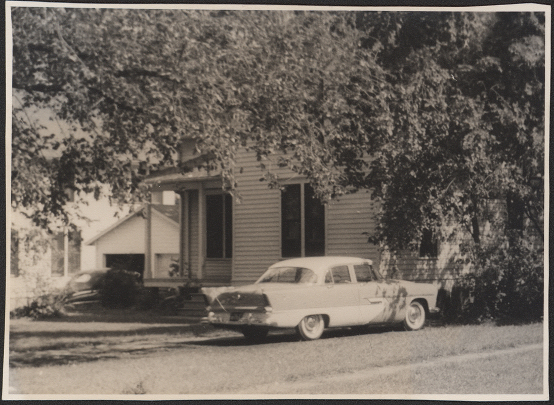 Photograph of Grounds Cottage. The photo depicts a house with a front porch and a car in the driveway. Much of the house is obscured by tree branches. A garage and part of another house are partially visible to the left. Caption reads "Built in 1882 as Laborer's Cottage for Jerry Sexton on site of Beach House moved west of Marsten Cottage. In 1924 it was moved to 2nd from East across from power plant."