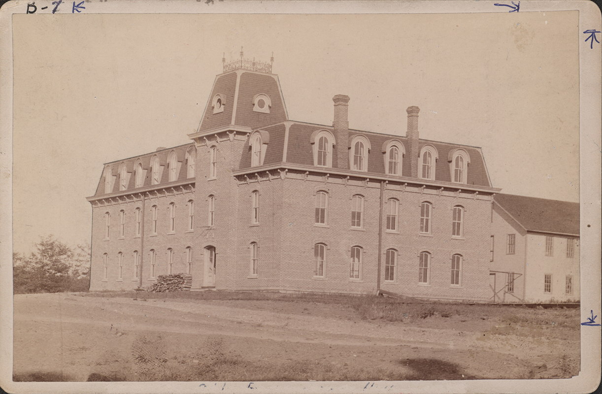 Photograph of the Laboratory of Mechanics. The photo depicts the front and side of the building. It is a brick building with three stories. A tower gable with a fence on top sits over the main entrance. A second building is partially visible to the right.