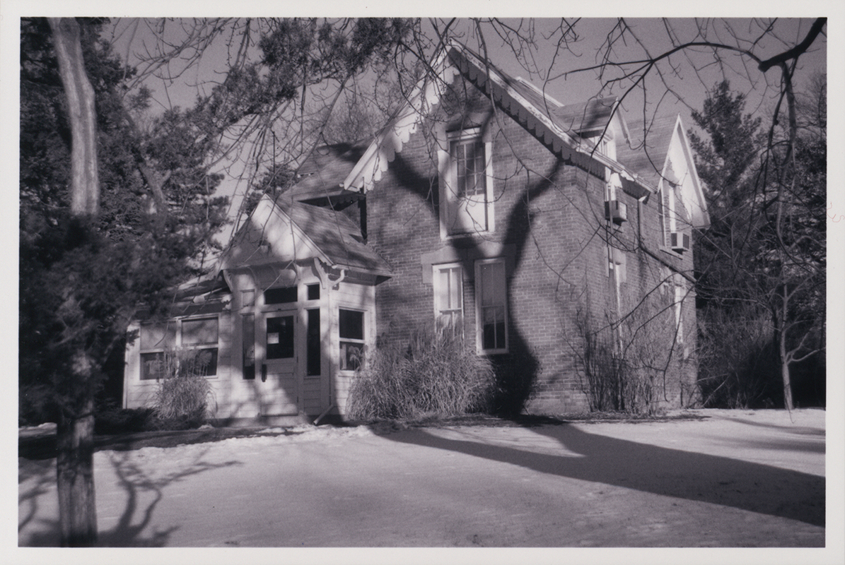 Photograph of Osborn Cottage looking north-west at the front door and east side of the building. The photograph shows trees casting shadows on the building and snow on the ground.