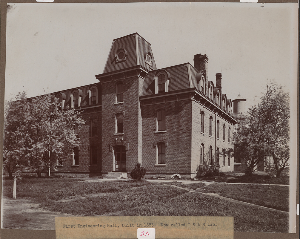 Photograph looking north-west of the southeast corner of the Laboratory of Mechanics. The Marston Water Tower can be seen to the north (right). Annotation: "First Engineering Hall, built in 1883. Now called T & A M Lab."