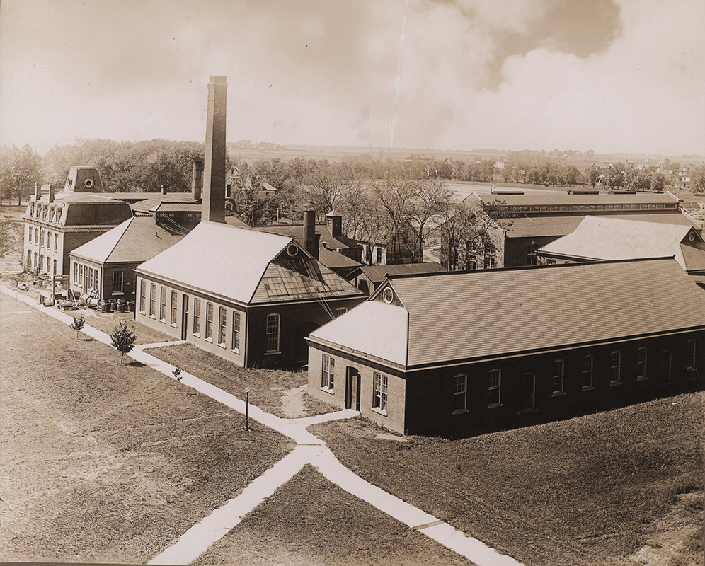 Photograph looking southwest at engineering laboratories. Following the sidewalk from left to right the buildings shown are the Laboratory of Mechanics, the Power Station, Building A, and the Aerospace Laboratory. To the west (right) of the Laboratory of Mechanics is the Quarantine Hospital, to the west of the Power Station is the Machine Shop, and to the west of Building A is Building B. Annotation: ''l. To r.; T. & A M Lab. (old Engineering Hall) 1884; site of M E Lab--picture of old power station--before 1908; Building A (old Foundry) 1898; Aeronautics Bldg (old Wood & Pattern Shop) 1898.''