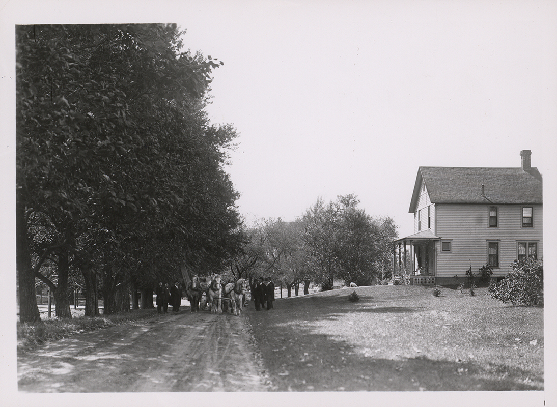 Photograph showing people and a team of horses standing in the street in front of the Engineer's Cottage. Annotation: ''Engineer's Cottage built in 1899 just east of the greenhouse. About 1915 it was moved to its present location as the eastern most cottage across from the heating plant. For some years it was the home of George Mitchell, farm superintendent.''