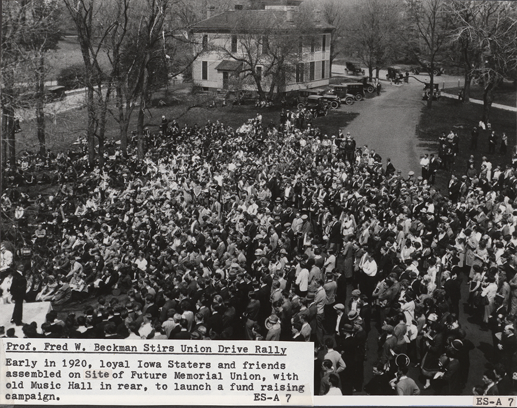 Photograph of Fred W. Beckman at the future site of the Memorial Union launching the fund raising drive for the Memorial Union. In the background cars are parked around Music Hall. Annotation: ''Prof. Fred W. Beckman stirs Union drive rally. Early in 1920, loyal Iowa Staters and friends assembled on site of future Memorial Union, with old Music Hall in rear, to launch a fund raising campaign.''
