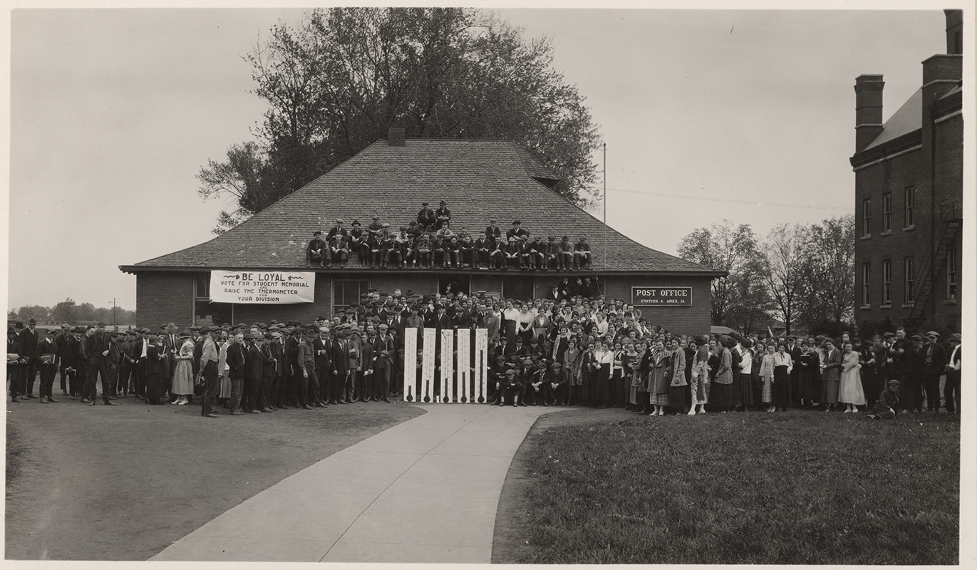 Photograph of people outside the Hub when it was the Post Office. There is a banner on the Hub reading ''Be loyal. Vote for student memorial and raise the thermometer for your division.'' Morrill Hall can be seen to the east (right).