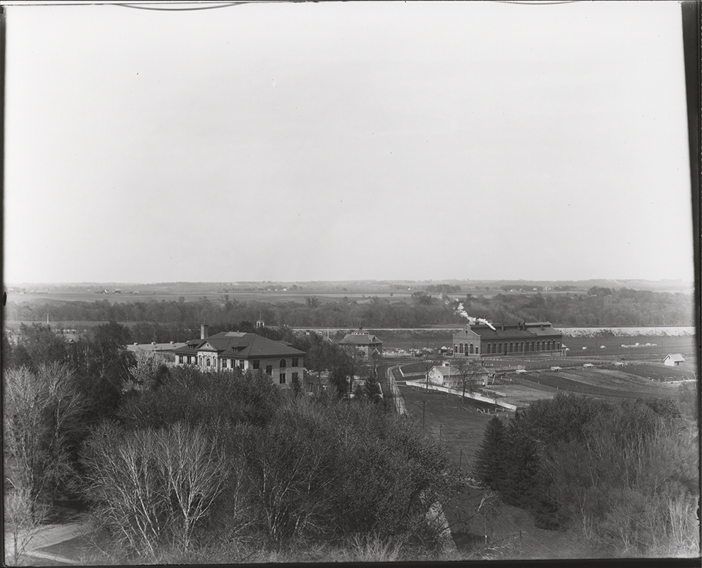 Photograph looking northeast past East Hall and down the Fort Dodge, Des Moines and Southern Railway tracks. In the distance a piece of the Carpenter Shop (Judging Pavilion #3) can be seen to the left of the tracks and the Power and Heating Plant can be seen behind where the tracks curve east. Annotation: ''East Hall, judging pavilion, power & heating plant, hog house. May 5, 1913.''