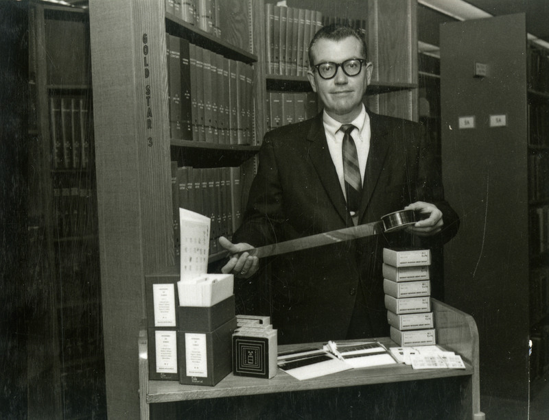 Warren B. Kuhn, Dean of the Library, is showing a reel of microfilm. Boxes of microfilm, an envelope of microfiche, and pam boxes with micro-opaques are displayed on the book truck.