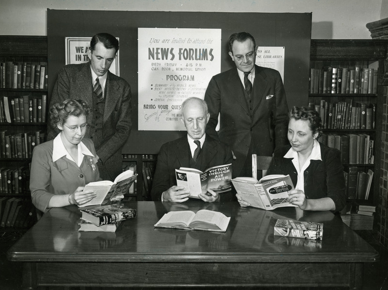 Five members of the library staff are readings books. Seated at the table (left to right) are Victoria Hardgrave, Charles H. Brown and Lucile Gib. Standing in the back row (left to right) are: Robert W. Orr and Keith Huntress.