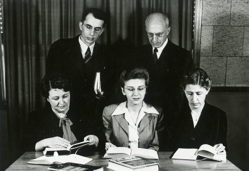 Three members of the library staff reading books are seated at a table, while two other library staff members are standing behind.