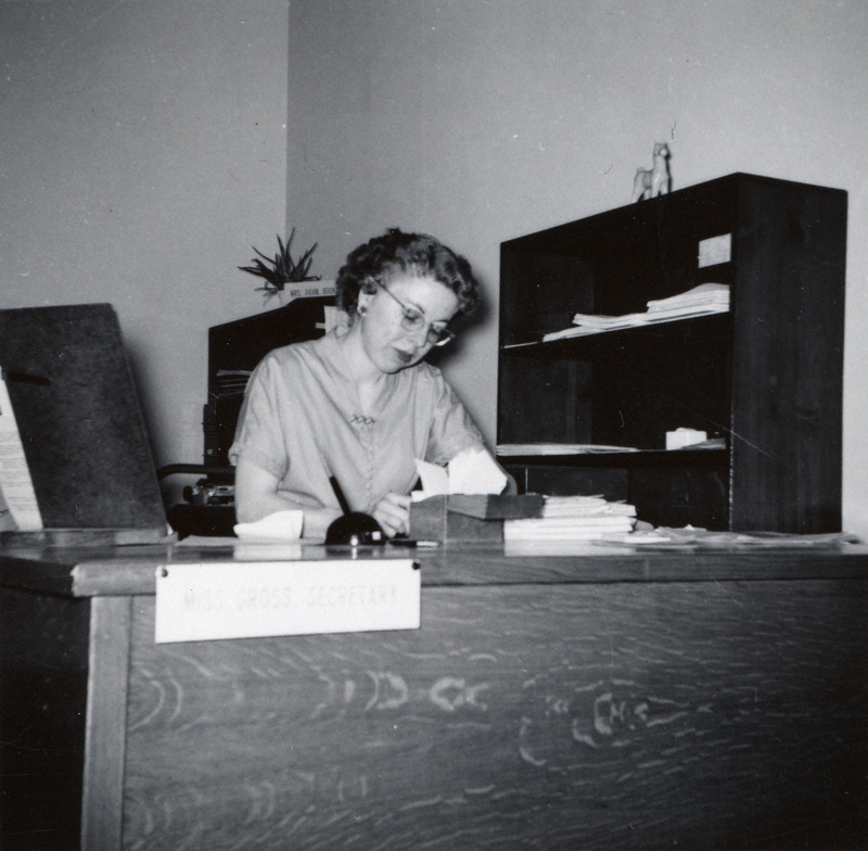 Margaret Mae Gross is seated at a desk in her office working on papers. A sign affixed to the desk front reads, "Miss Gross, Secretary.".