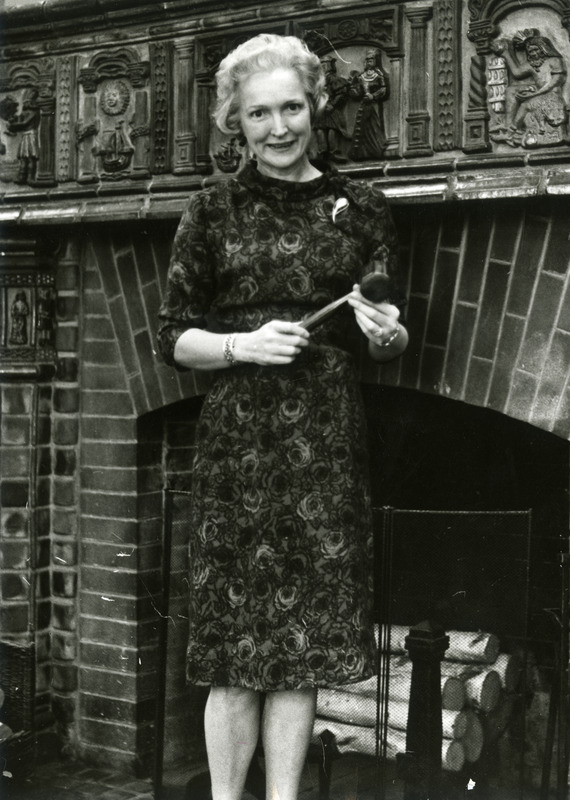 Dorothy Kehlenbeck in front of fireplace in Room 105 holding a gavel made from a wooden water main of old Chemistry Hall which was unearthed when Pearson Hall was being built. Professor Breckinridge made the gavel.