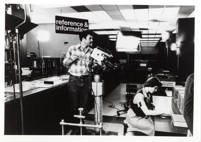 One person is using a video camera to record another person who is seated at a desk.