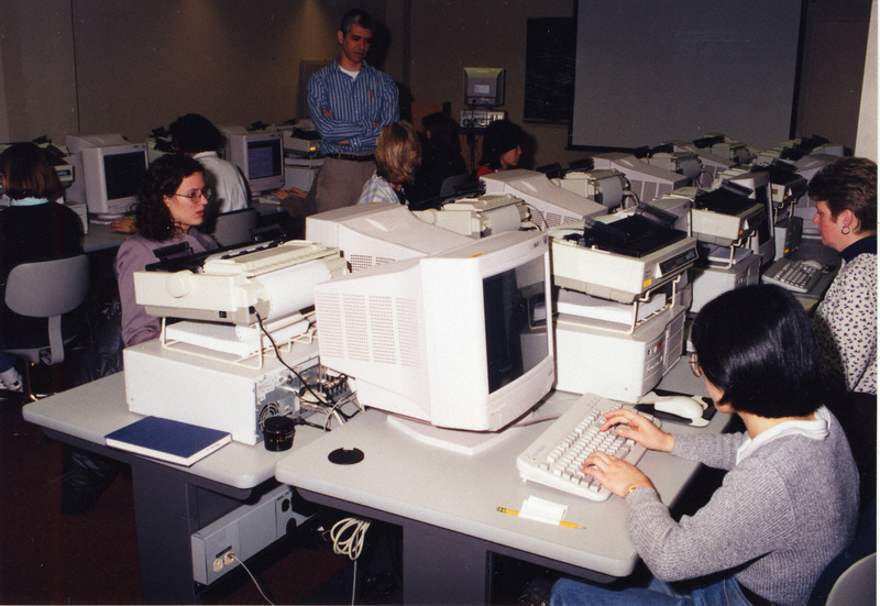 People are seated at a double bank of computer terminals in the library instruction lab. An instructor standing near by instructing the students.