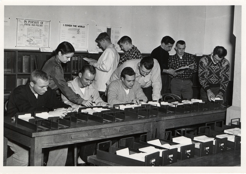 A group of people are being instructed in library card catalog records, Some of the people, seated at a table are examining individual cataloging cards in drawers. Other standing individuals are consulting books.