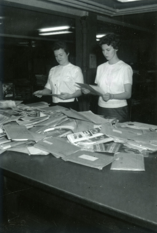 Cathy Cormack and Linda Mandrup stand at a large table covered with envelopes and other incoming mail. They are in the process of sorting the mail so it can be delivered to various departments in the library.