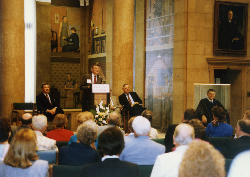 Former ISU president Gordon Eaton is at the podium speaking at the dedication of the Presidential Portrait Gallery in the library's upper rotunda lobby. Martin Jischke is sitting on the left, and the presidential portrait of Gordon Eaton is displayed on an easel on the right.