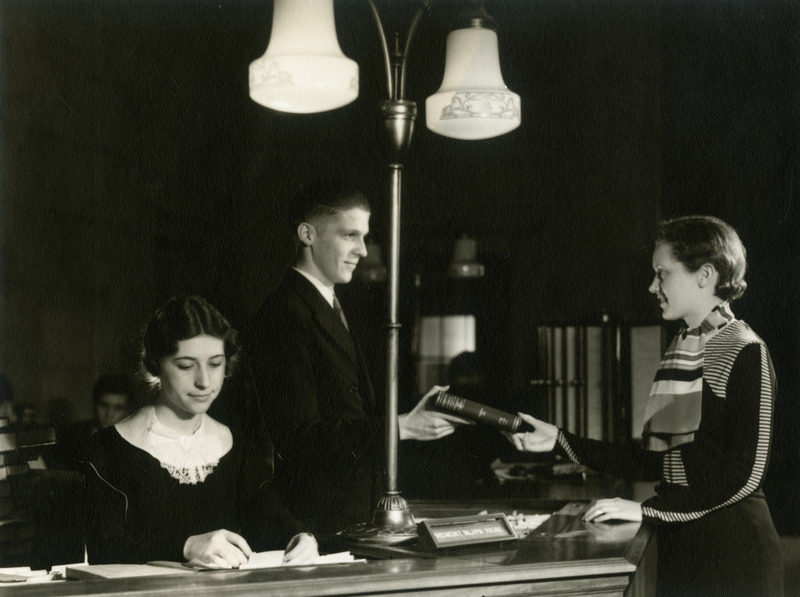 At the Loan Desk (now called Circulation Desk), a male staff member hands a book to a female student, while a female staff member sits nearby.