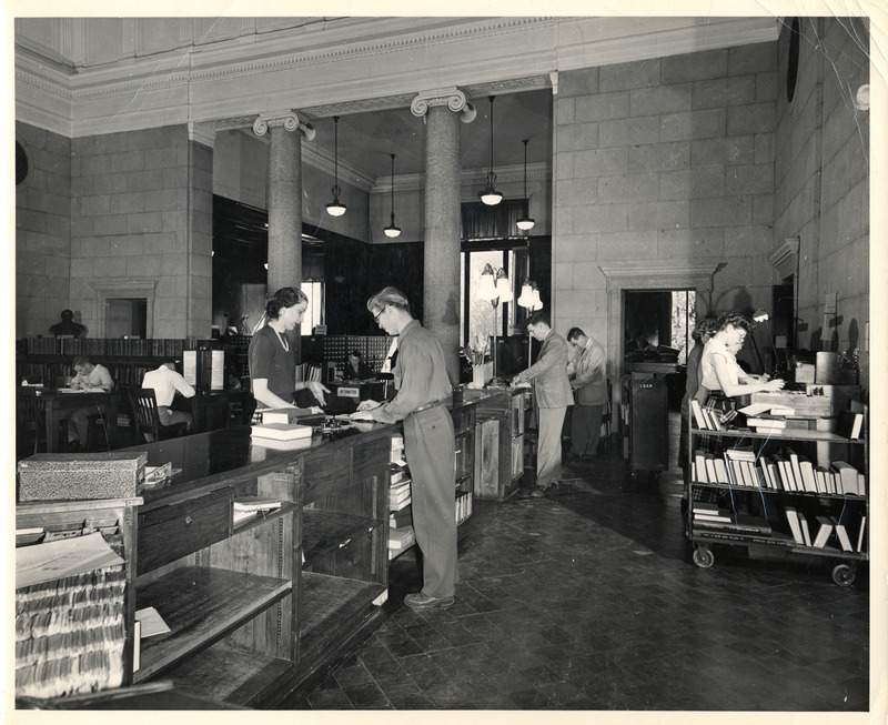 The Library Loan Desk (now called Circulation Desk) is viewed from the librarian's side, with a male staff member helping a female patron while other staff work with card files and book trucks behind the desk.