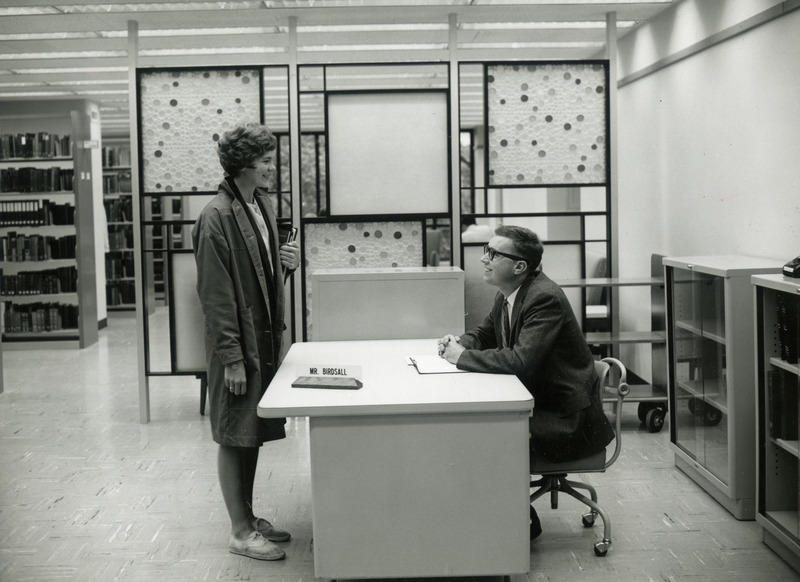 A female student stands and chats with reference librarian William Birdsall, seated at a metal desk next to a room divider with multi-colored circles, circa 1961. There were reference areas like this on each of the first three floors after open stacks were adopted with the opening of the 1st Addition.