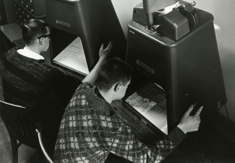 Two male students read microfilmed newspapers on Recordak microfilm readers in the library.