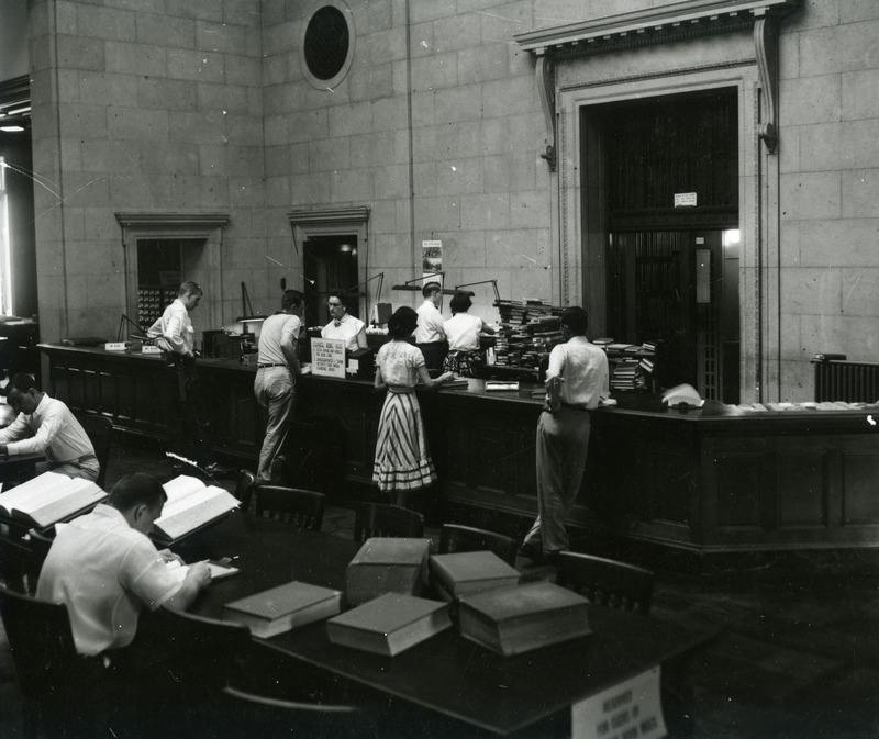 A library staff member helps four patrons lining up at the Loan Desk (now called the Circulation Desk), while two staff members work near piles of books behind the desk. A sign on the desk explains procedures for charging books.