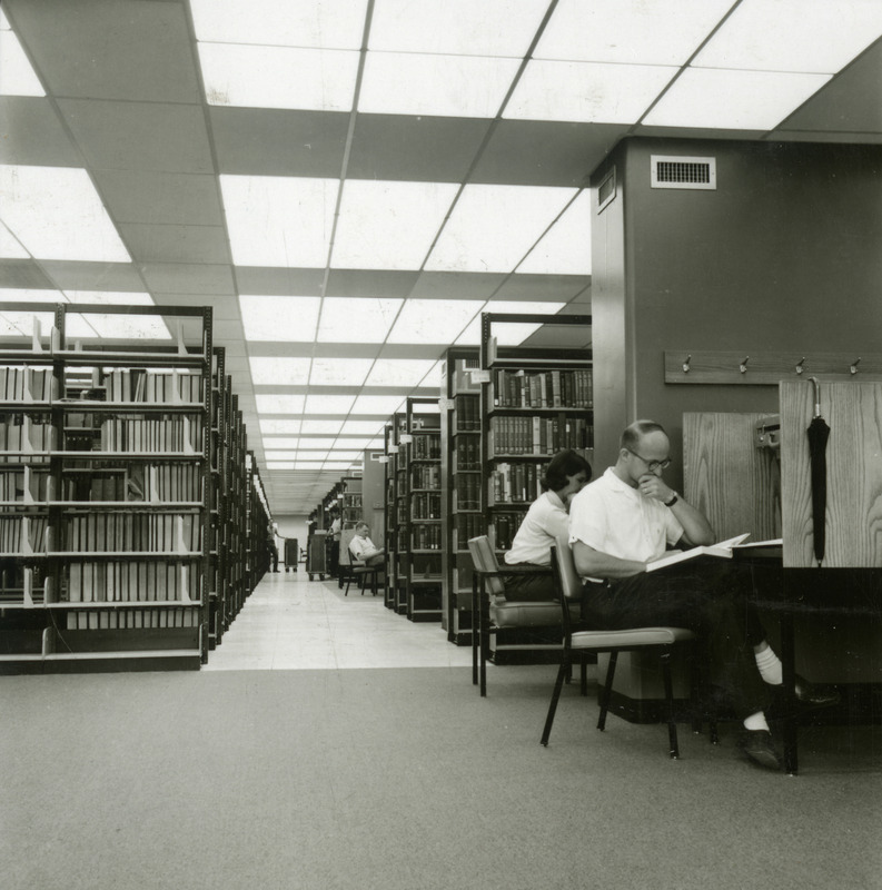 Three library patrons read at study carrels while workers shelve books in the stacks of the newly completed 2nd Addition to the library.