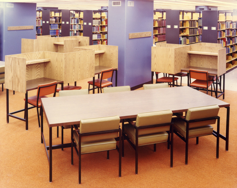 Library study tables and desks sit on orange carpet next to blue columns in the 1st Addition.