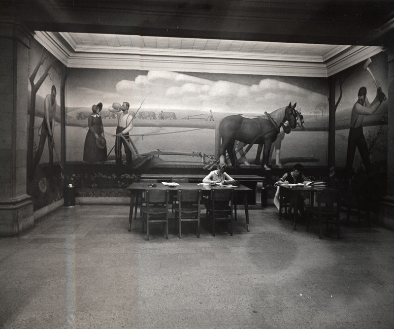 Two female students study at tables in front of the Grant Wood mural "Breaking the Prairie.".