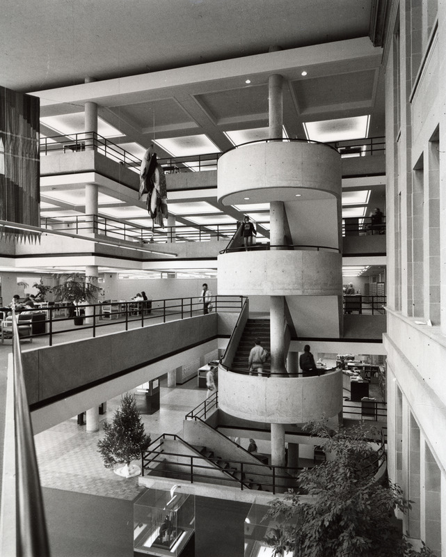 All four floors are visible in this view of the circular staircase in the 3rd Addition, taken from the second floor bridge. The fish soft sculpture and a banner hang on the left. On the first floor, there is a decorated Christmas tree and glass display cubes, and the card catalogs and reference desk are slightly visible.