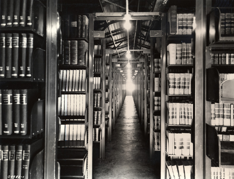 A dark aisle through the middle of the old library storage building is lit by overhead light bulbs, illuminating the concrete floor and little-used volumes filling the tall metal shelves.