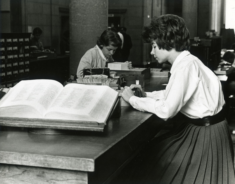 Two young women on either side of a tall table look through drawers of the card catalog in the upper rotunda lobby.