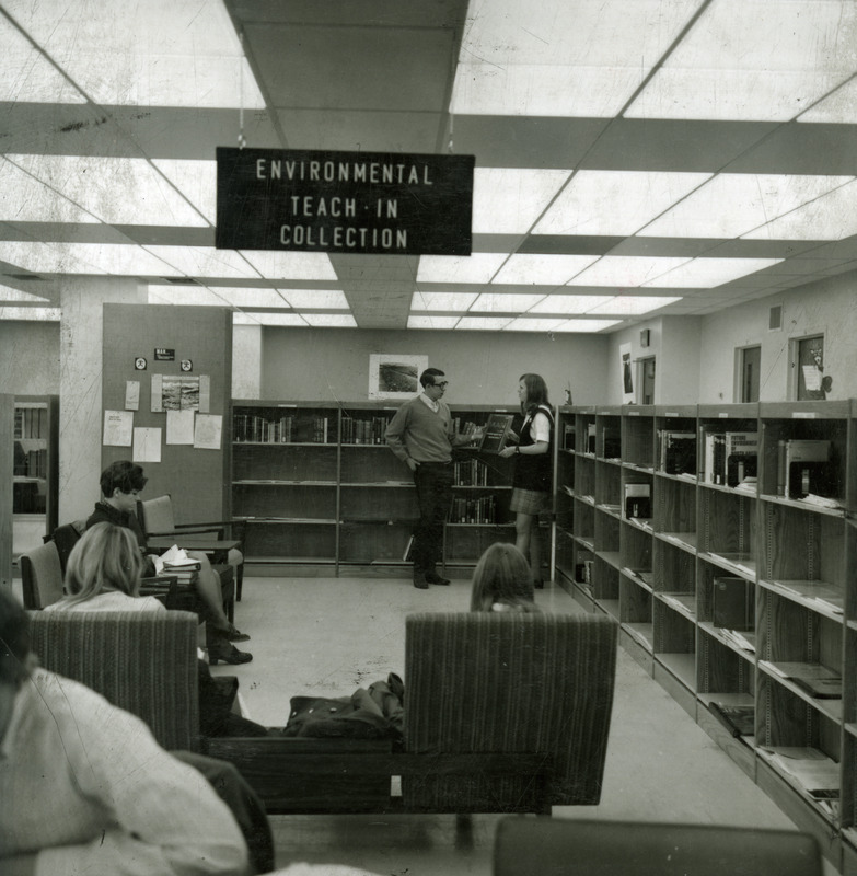 Two students hold a book entitled "Man: An Endangered Species?" amid the low shelves of the Environmental Teach-In Collection, while other students sit in the chairs near the collection.