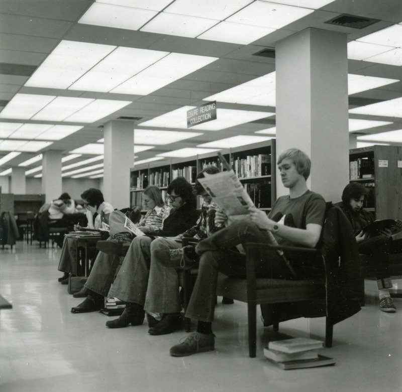 Students read newspapers while sitting in chairs by the Leisure Reading Collection.