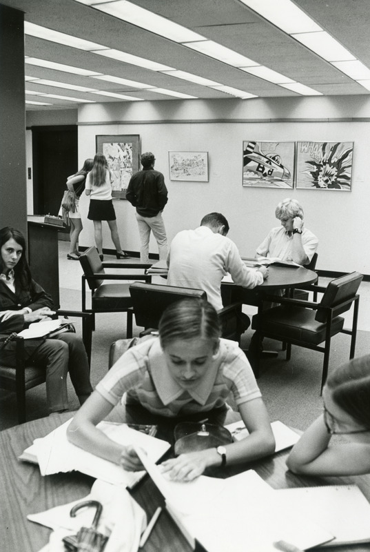 Students study at tables while others view art on the wall in the background.