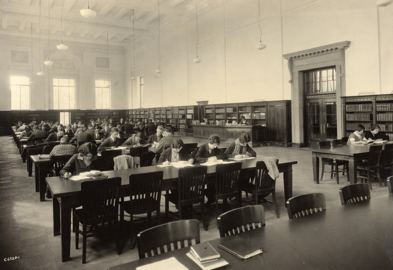 Students are studying in the Main Reading Room (currently called the Periodical Room) in the library. Two library staff members are working at the desk.