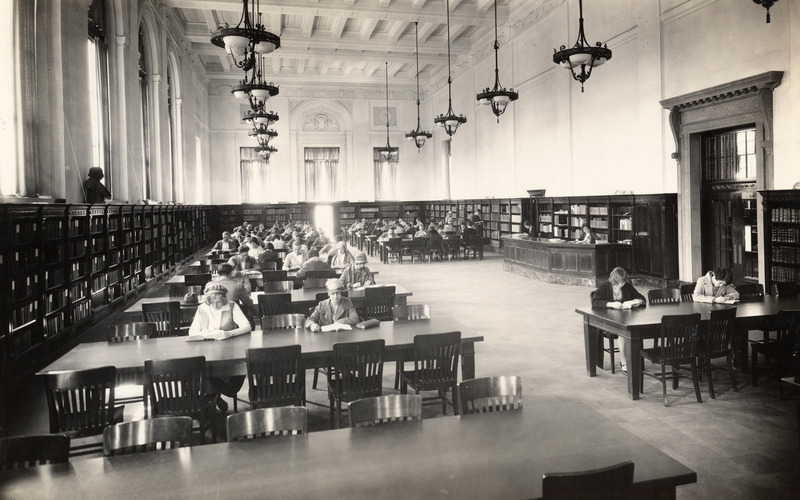 Students are studying in the south end of the Main Reading Room (currently called the Periodical Room) in the library. Some of the women have hats and coats on. Two library staff members are working at the desk.