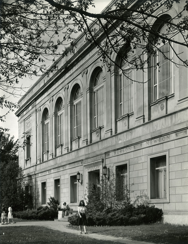 A woman is seated on the bench as two women approach the east entrance to the library. Another women is walking down the path from the library.