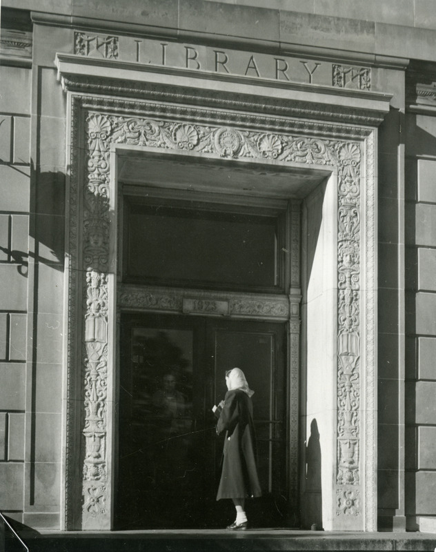 A woman dressed in a scarf and coat is entering the east door of the library as another person, partially visible, is exiting the library.