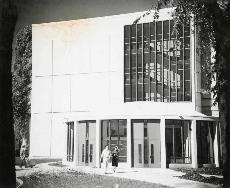 A couple is walking past the rotunda on the south side of the 1st library addition. Another man is wheeling a cart toward the entrance.