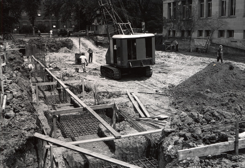 Workmen have started work on the foundation for the 1st addition. Vertical rebars are sticking out of the foundation. A workman is operating a crane owned by Thomas Construction Company to lift a drum-shaped object. The workmen's' office, two vehicles and Gilman Hall are visible in the background.