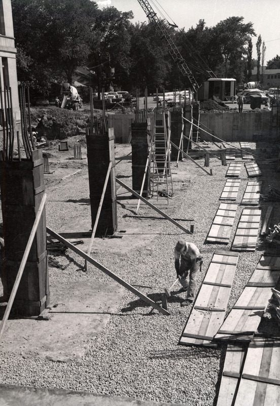 This below ground level view shows several rebar-reinforced columns. A workman is pounding in a wood reinforcement for the column. Two cement mixers and a crane are parked in the above ground parking lot.