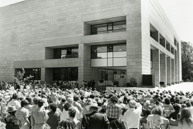 The university community is gathered outside for the dedication of the 3rd addition to the library. A man is speaking at the podium. To the right of the podium is the covered portrait of Dr. Robert Parks which was unveiled at the ceremony.