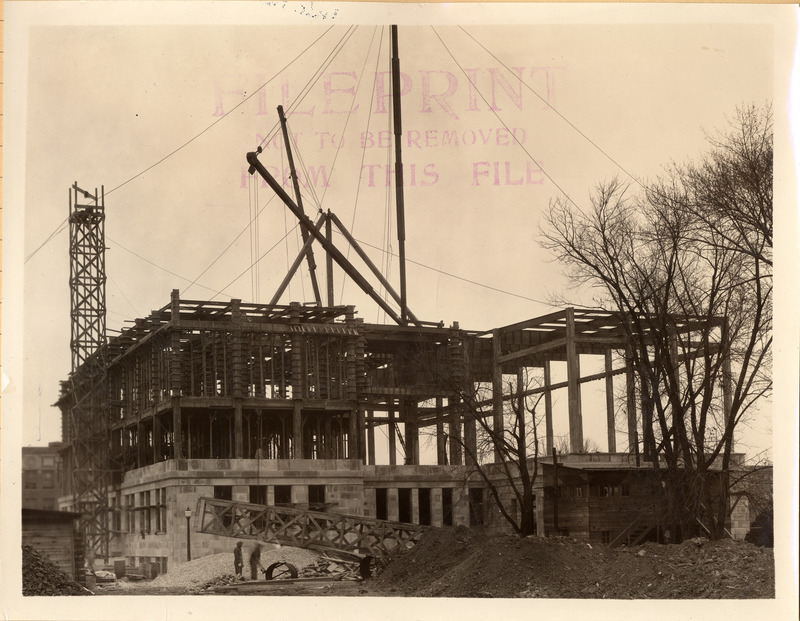 Construction work continues on the original library building. The exterior of the first floor is taking shape while the upper floors are still framed. A construction office appears in the lower right corner.