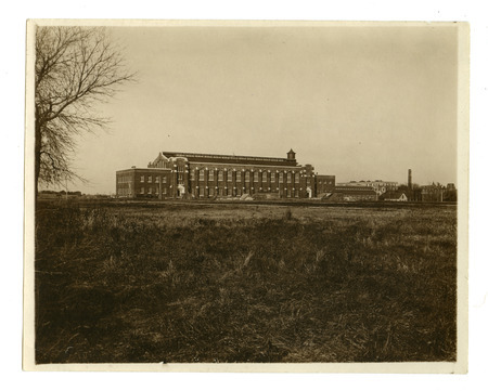 Viewed from a distance, the back (south) side of State Gymnasium is visible. Lab of Mechanics, Machine Shop, quarantine hospital and Beardshear Hall can be seen on the far right. The Marston Water Tower is in the background. Building supplies are laying around the building.