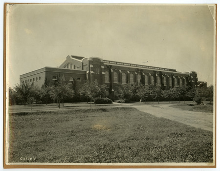 The front of the State Gymnasium is viewed from northeast with trees hiding part of the building. The fence of an athletic field is on the right with people sitting in the corner, and a dirt road leads past it towards the building, in this darker exposure.