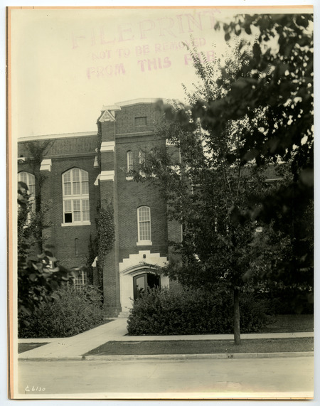 The front (north) entrance on the west end of the State Gymnasium is marked by a tower, with an open door on the right and an open window on the upper left. Trees partially obscure the building, and ivy climbs the left side of the tower.