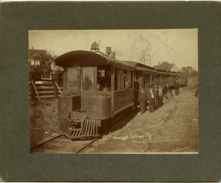 Frank Lang, motorman for the Ames & College Railway (The Dinkey) is seated in the locomotive while other individuals stand beside the train. The third and fourth standing individuals from the left are identified as M. K. Smith, manager and Hank Wilkerson. The other persons are not identified. Writing on the front of the photograph suggests that it was taken during the Excursion Days, October 5-6, 1906.