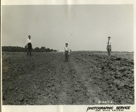 A group of three men are standing in a plowed field. The furrow in the middle is considerably lower than those on either side.