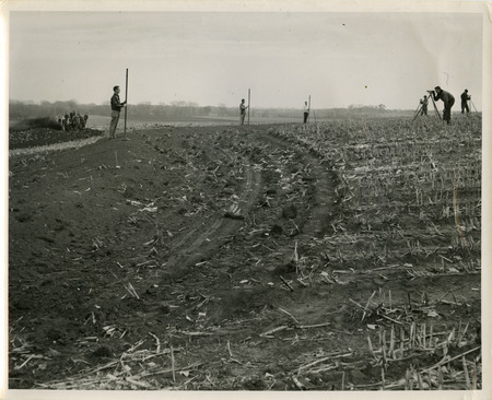 Several individuals are using survey rods and transits to build terraces in a farm field. Another group of individuals and a tractor are in the background.