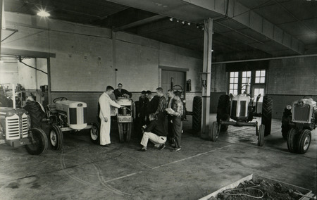 An instructor explains the front mechanics of a tractor to six students in a laboratory with five other tractors.